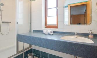 A modern bathroom with a shower and a large sink. On the shelf are two rolled towels and a mirror can be seen in the background.