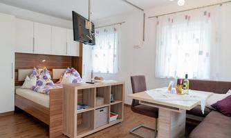 A modern room with a double bed and a television. The room is bright and cozily furnished, with a dining table and a comfortable couch.