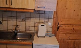 A small kitchen with wooden cabinets and a sink. Next to the sink is a dishwasher and a wooden door is visible in the background.
