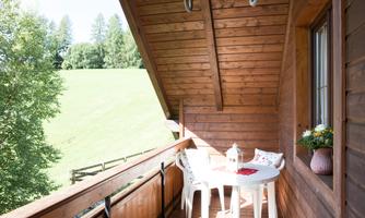 A beautiful balcony with a round table and white chairs. The surroundings are green and the sun is shining.