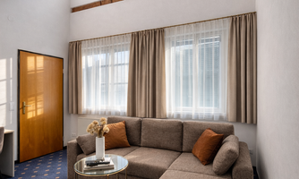 A cozy living room with a bright sofa and round table. Large windows let in plenty of light, and the ceiling has visible wooden beams.