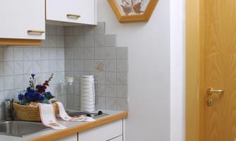 A small, bright kitchen with white cabinets and a sink. A decorative picture hangs on the wall, and the door is made of wood.