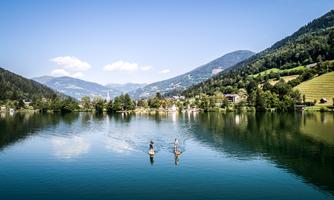 Two people are standing on stand-up paddleboards on a calm lake. In the background, mountains and green meadows can be seen under a clear blue sky.
