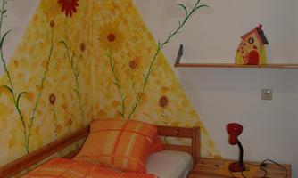 A cozy bedroom with a single bed and a colorful wall painting of flowers. A red lamp sits on the nightstand and there is a small wooden wardrobe.