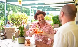 Two men are raising their glasses to toast in a sunny outdoor area of a restaurant. In the background, plants and other guests can be seen.