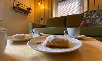 A cozy room with a green couch and warm wooden paneling. On a table, there are plates with pastries and cups, ready for coffee or tea.