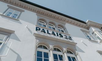 An elegant building with the inscription "PALAIS 26". It is painted white and has large windows.