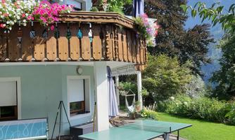 A beautiful house with a balcony, surrounded by colorful flowers. In the foreground, there is a table tennis table on a green lawn.