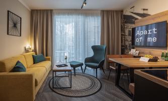 A modern apartment with a cozy yellow couch and a round rug. There is a dining table with chairs and large windows that let in plenty of light.