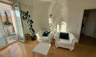 A bright room with two white armchairs and a small table. Light falls on the floor through an open window, and a plant stands in the corner.