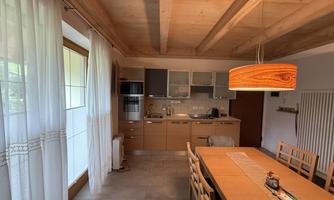 A modern kitchen with a wooden ceiling and a large dining table. The room is bright and friendly, with large windows and a warm light source.