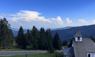 A picturesque mountain landscape with green forests and a small church in the foreground. The sky is clear and shines in various shades of blue.