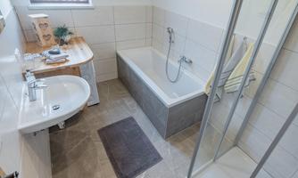 A modern bathroom with a bathtub, a shower, and a sink. The walls are light tiled, and there is a wooden shelf with decorations.