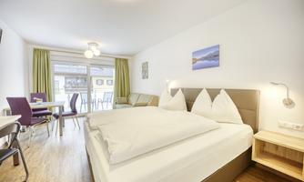 A modern hotel room with a large bed and a cozy seating area. Daylight streams through the windows, and a beautiful view of the terrace is visible.