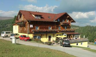 A well-kept inn in the mountains with flower balconies. Parking spaces and a beautiful landscape surround the building.