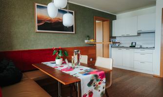 A cozy dining area with a table, decorated with a colorful tablecloth and plants. In the background, there is a modern kitchen and a piece of artwork on the wall.