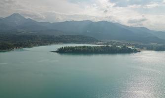 A tranquil landscape with a turquoise blue lake and a small island. In the background, gentle mountains and a cloudy sky can be seen.