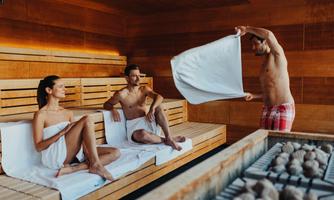 A sauna with three people relaxing. A man is waving a towel while the other two are sitting on the bench.