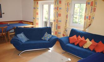 A cozy living room with blue sofas and colorful cushions. Large windows let in plenty of light, and there is a dining table in the background.
