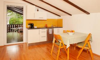 A modern kitchen with wooden flooring and a dining table for four people. A sunny balcony is visible through the glass door.