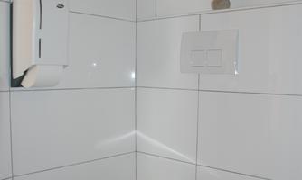 A modern bathroom with white tiled walls. The toilet is located in the corner, next to it is a paper towel dispenser.