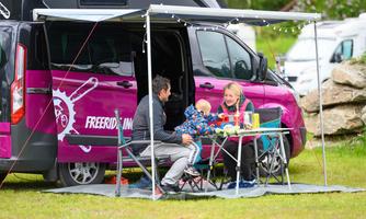 A family sits in a camping area in front of a motorhome. They enjoy a meal under a sunshade.