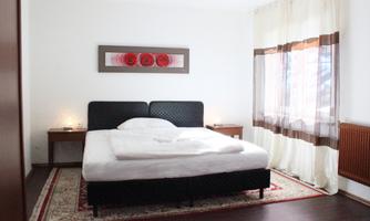 A modern bedroom with a large bed and stylish nightstands. Large windows with bright curtains provide plenty of daylight.