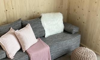 A cozy living room with a gray sofa, colorful cushions, and a soft throw. Decorative pictures hang on the wall, and there are round poufs in natural colors.