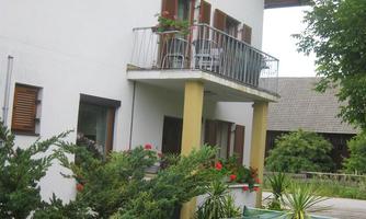 A two-story house with a balcony and blooming plants. In the foreground, there is a table with chairs on a paved area.