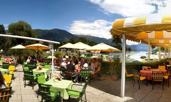 An inviting outdoor restaurant with colorful tables and umbrellas. In the background, mountains and a lake can be seen as guests enjoy the beautiful weather.