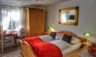 A cozy bedroom with a double bed and red pillows. There are two chairs at a small table, and the room has large windows with white curtains.