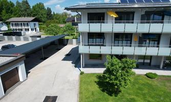 A modern building with balconies and a well-maintained garden. In the background, there are other houses and a parking lot.