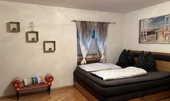 A cozy bedroom with a large bed and decorative pillows. The room has wooden floorboards and a windowsill with curtains.