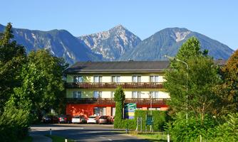 A modern hotel with a green facade is located on a quiet street. In the background, majestic mountains rise under a clear sky.