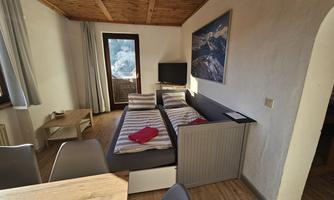 A cozy living space with a bed, a television, and a wooden table. The large windows offer a view of the alpine landscape.