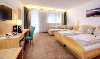 A modern hotel room with two beds, a sofa, and a television. Large windows provide plenty of daylight.