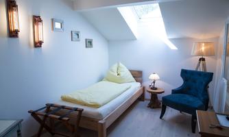 A bright room with a single bed and a cozy blue armchair. The wall has picture frames and a window lets in daylight.