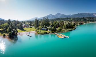 A quiet lake with clear, greenish water surrounded by mountains. On the other side, loungers and recreational areas can be seen.
