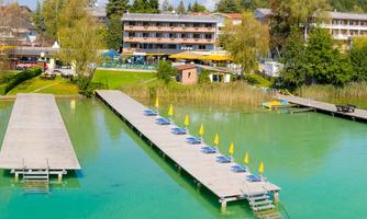 A beautiful lake with a wooden dock and loungers under yellow sun umbrellas. In the background, trees and a hotel can be seen.