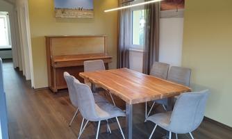 A modern dining area with a wooden table and six chairs. In the background, there is a piano and large windows let in plenty of light.