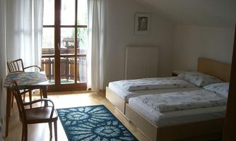 A simple room with two beds, a table, and a chair. Large windows let in plenty of light and provide a view of the balcony.