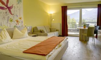 A modern hotel room with a large bed, a sofa, and a dining area. The windows offer a view of the mountains and plenty of natural light.