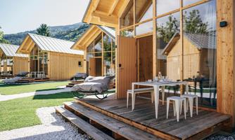 Modern wooden cabins with large windows in a green setting. The outdoor area features a terrace with a table and chairs.