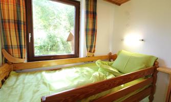 A cozy bedroom with a wooden bed and fresh, green bedding. A large window lets in plenty of daylight and offers a view of the greenery.
