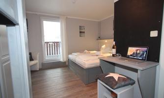 A modern room with a double bed and a desk. There is a window with a balcony view and a bright, friendly atmosphere.