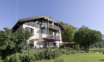 A modern house with multiple floors in the midst of a well-maintained garden. The sky is clear and blue, surrounded by trees and green grass.