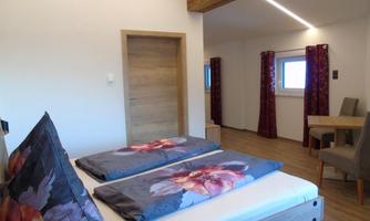 A cozy bedroom with two beds and colorful cushions. The room is bright and modernly furnished, with a small table and chairs.