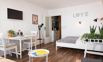 A bright and modern apartment with a bed, a table, and plants. The word "LOVE" is on the wall, and a telescope is set up.