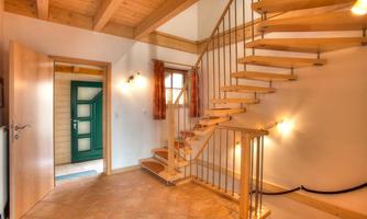 A bright hallway with a modern wooden staircase and large windows. The open door leads to a green entrance area.