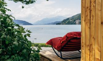 A cozy lounge chair with a red cushion is placed on a wooden terrace. In the background, a calm lake and the surrounding mountains can be seen.